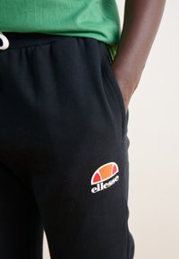 Person i svarta sweatshirtbyxor med Ellesse-logga på låret och grön tröja, hand delvis i fickan mot neutral bakgrund.