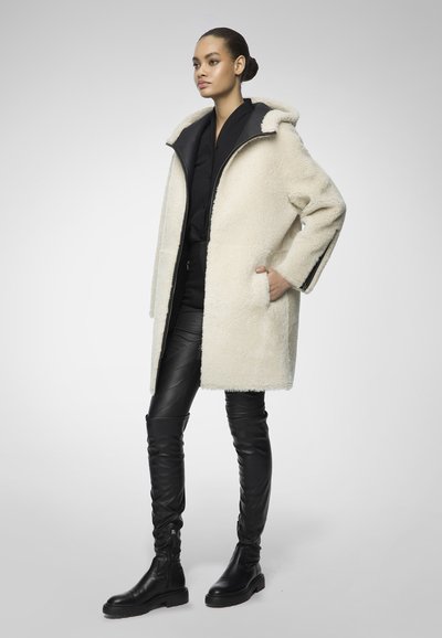 Donna in piedi con le mani in tasca, indossa un cappotto bianco in pelliccia, una blusa nera, pantaloni in pelle neri e stivali neri fino al ginocchio.