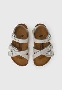 Hõbedased sädelevad väikelapse sandaalid reguleeritavate pandalde ja pruuni kujundatud jalataldadega, esitatud valgel taustal.