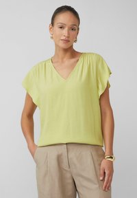 V-hals blouse met korte mouwen van licht limoengroene stof, met zachte plooien en een ontspannen pasvorm; gecombineerd met beige broeken en een gouden armband.