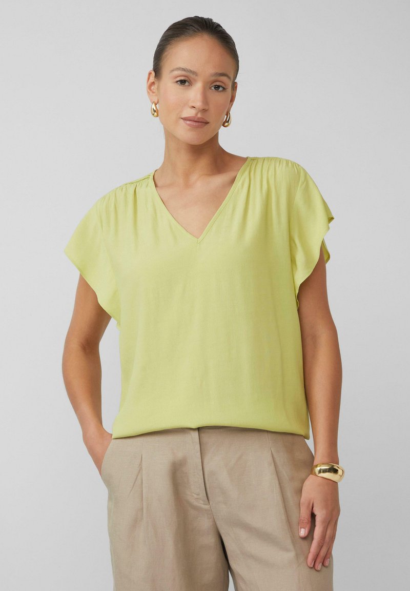 V-hals blouse met korte mouwen van licht limoengroene stof, met zachte plooien en een ontspannen pasvorm; gecombineerd met beige broeken en een gouden armband.