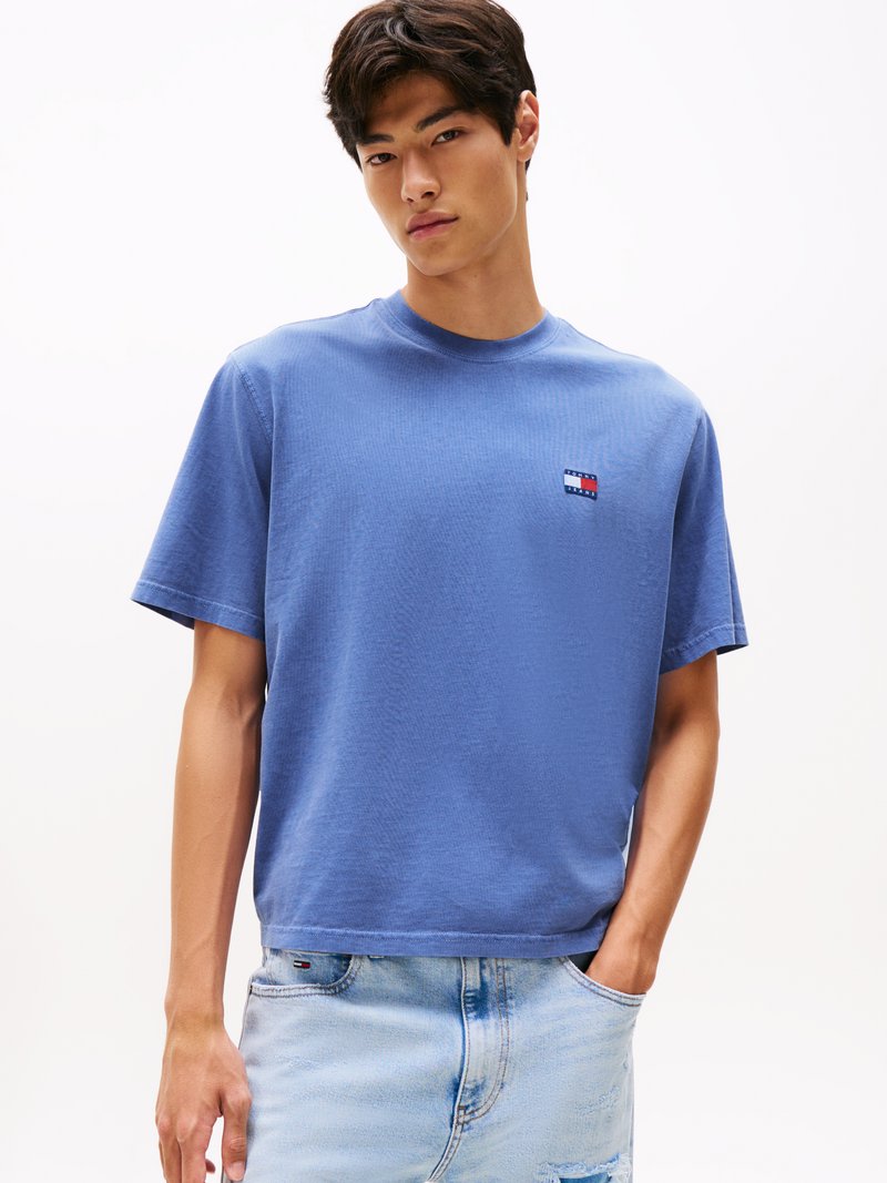 T-shirt blu in cotone a maniche corte con una texture a coste e una piccola toppa con logo sul petto sinistro. Indossato con jeans chiari strappati.