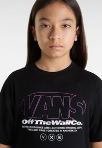 Sort bomulds t-shirt med lilla og hvide "VANS Off The Wall Co." grafiske tekst. Korte ærmer og rund halsudskæring design.