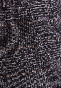 Gros plan sur un tissu à carreaux gris et marron avec une étiquette de marque en métal noir "GABBIANO" cousue près de la couture d'une poche.