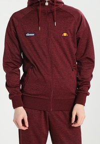 Burgundy zip-up hoodie with a textured fabric, featuring a logo patch, hood with drawstrings, and two front zippered pockets.