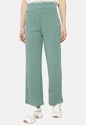 Pantaloni larghi verde chiaro con una texture liscia, caratterizzati da un alto punto vita e due cuciture frontali, indossati con sneakers bianche.