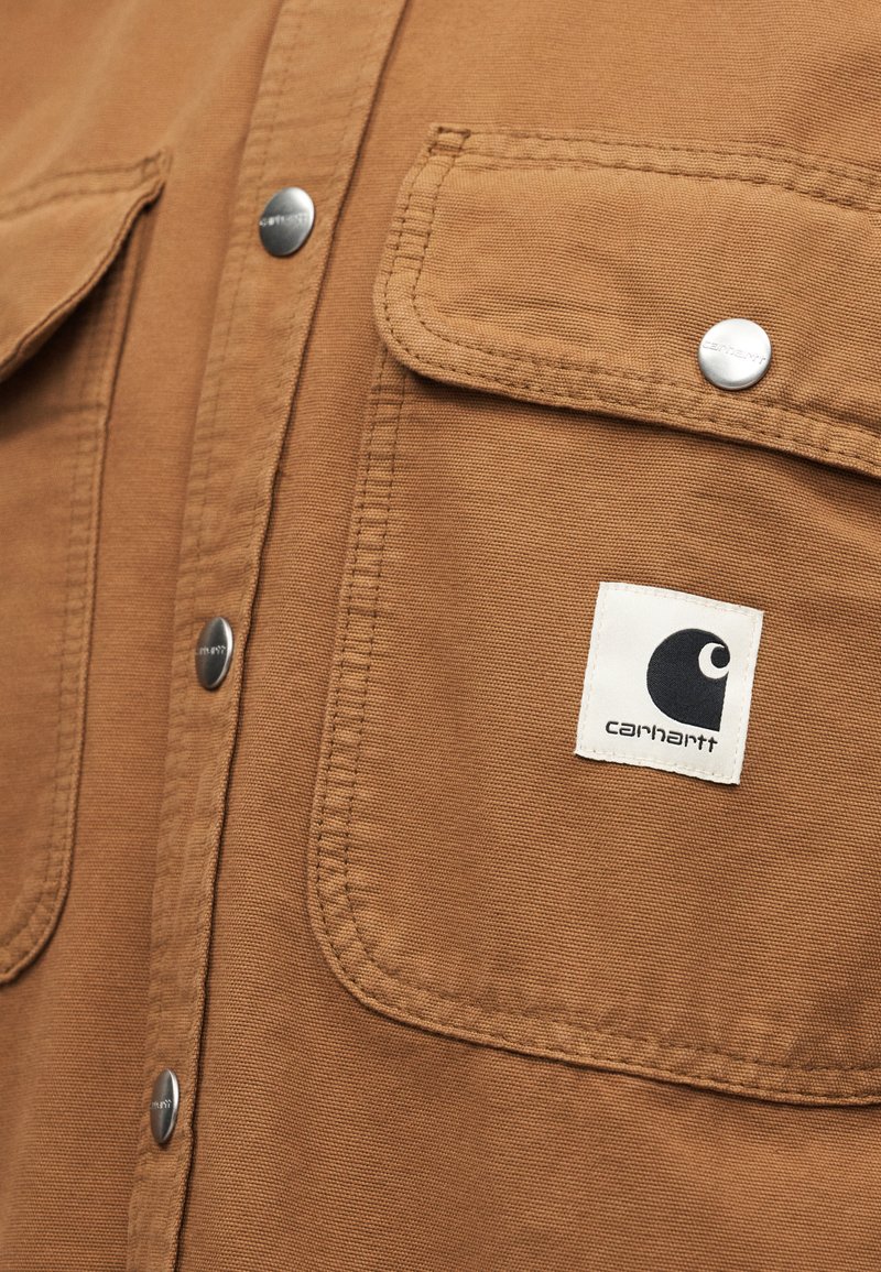 Tasca con patta e bottone a pressione in metallo su camicia in tessuto marrone, con una toppa rettangolare bianca con il logo Carhartt.