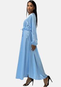 Abito maxi in satin blu chiaro, con maniche lunghe, vita sagomata e cordoncino. Texture liscia e silhouette fluida. Tacchi alti neri.