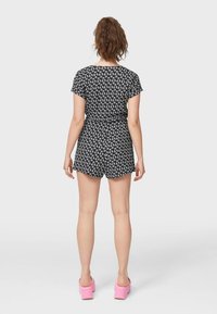 Sort jumpsuit med blomsterdesign, korte ærmer og shorts. Har en tætsiddende talje og løs pasform. Båret med lyserøde platformssko.