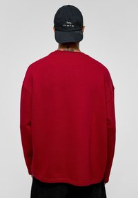 Rode sweatshirt met lange mouwen, een relaxte pasvorm en een ronde hals, gecombineerd met een zwarte pet met witte borduursel aan de voorkant.