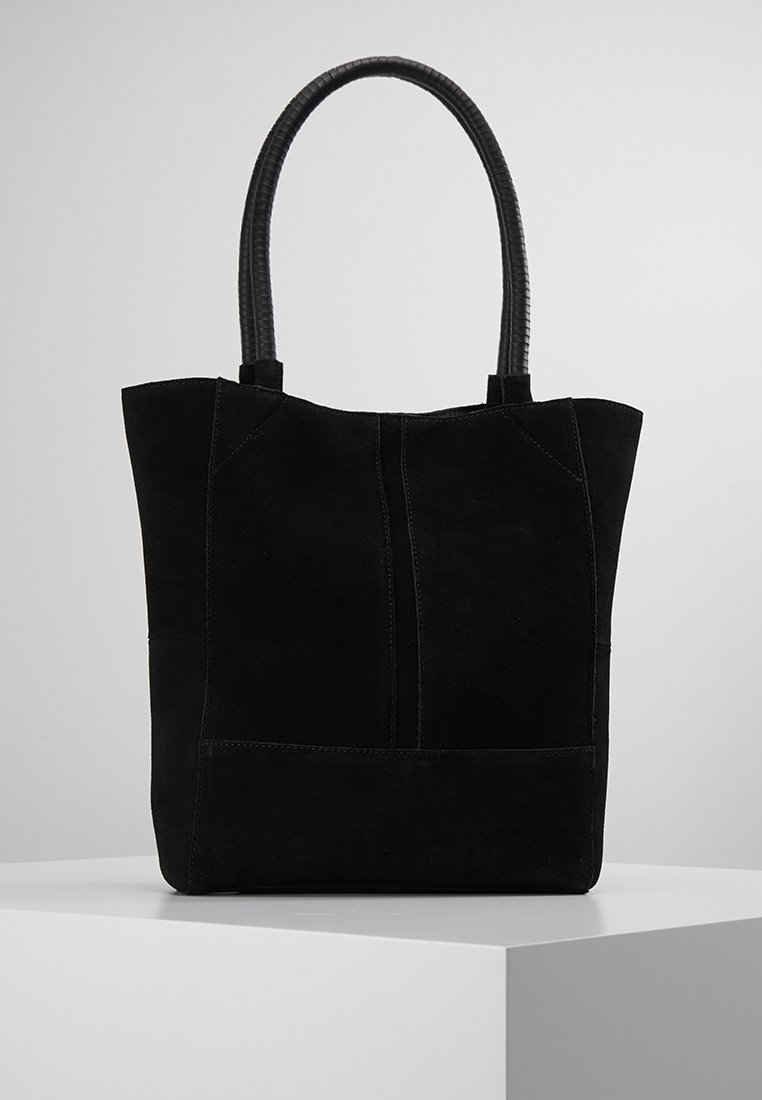 Sac cabas en daim noir avec poignées arrondies noires texturées, détail de couture verticale, exposé sur un socle blanc sur fond gris.