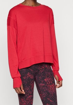 Persoon draagt een losse rode trui met lange mouwen en duimgaten en zwarte leggings met rode abstracte patronen.
