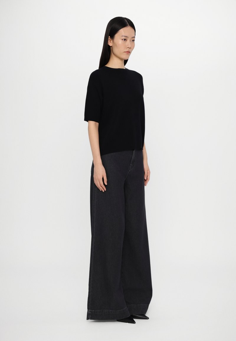 Black short-sleeve top paired with wide-leg dark denim jeans. Model stands sideways, showcasing the relaxed fit and elongated silhouette.
