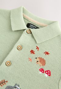 Polo vert clair en maille avec un col côtelé, des boutons en bois, et des broderies d'un hérisson, de champignons et de feuilles colorées.