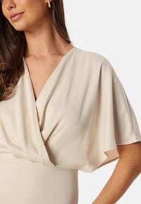 Beige omslagstopp med V-ringning och draperade ärmar, tillverkad av mjukt tyg. Den anmärkningsvärda mjuka texturen och det eleganta designen förstärker dess enkelhet.