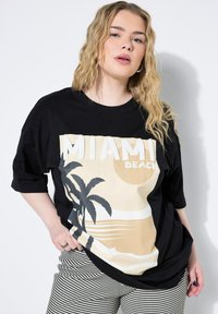 Mustapohjainen oversize t-paita, jossa on beige Miami Beach -grafiikka palmupuiden siluettien ja aaltojen kanssa. Yhdistetty raidallisiin, tekstitettyihin housuihin.
