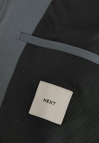 Tissu noir texturé avec bordure grise et une étiquette blanche cousue avec l'inscription « NEXT » en lettres noires.