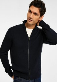 Marineblauw geribd gebreid vest met hoge kraag, lange mouwen en ritssluiting. Gecombineerd met blauwe spijkerbroeken.