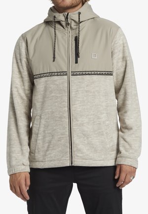 Veste à capuche gris clair et beige avec un tissu texturé, des accents noirs, des poches zippées et des cordons ajustables à la capuche.