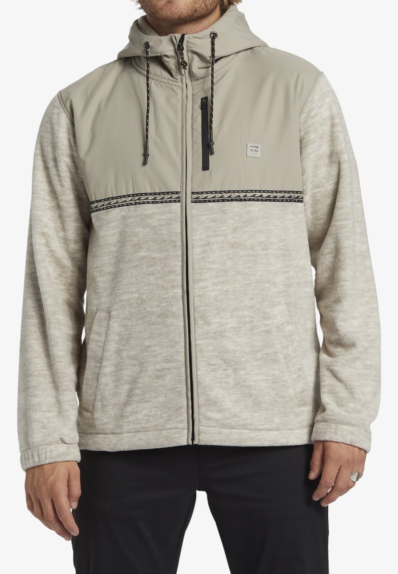 Lichtgrijze en beige hooded jack van textuurstof, met zwarte accenten, ritszakken en verstelbare koorden bij de capuchon.
