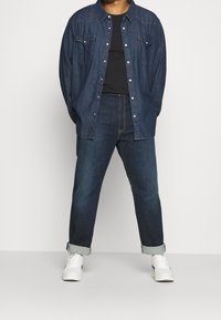 Džínová košile tmavě modré barvy se slim fit střih, zapínáním na knoflíky vpředu, dvěma náprsními kapsami a dlouhými rukávy, vrstvená over černým tričkem a džínami.