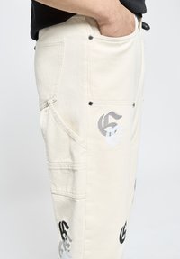 Pantalones cargo de algodón beige con logotipos bordados en negro y gris, que presentan múltiples bolsillos y detalles en hardware plateado.