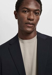 Blazer bleumarin cu revere croite, asortat cu un pulover tricotat de un bej deschis. Textură de stofă netedă, croi clasic și design minimalist.