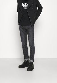 Man som bär en svart motorcykeljacka, svart grafisk t-shirt, mörkgrå uppvikta jeans och svarta läderskor med snörning, stående mot vit bakgrund.