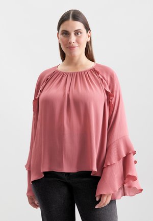 Blusa rosa pallido con texture plissettata, scollatura rotonda e maniche a volantino, abbinata a jeans grigio scuro.