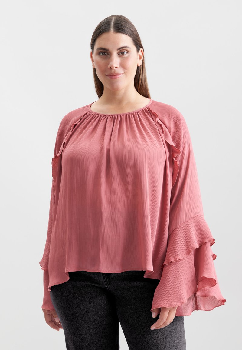 Blush-pink Bluse mit plissierter Textur, rundem Ausschnitt und Rüschensäumen, kombiniert mit dunkelgrauen Jeans.
