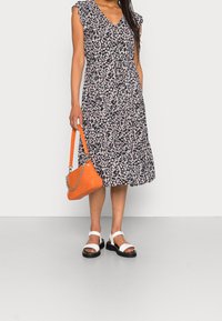 Kleid mit floralem Druck auf schwarzem Hintergrund, mit Rüschensleeves, tailliertem Design und gestuftem Rock. Kombiniert mit weißen Sandalen und einer orangefarbenen Handtasche.