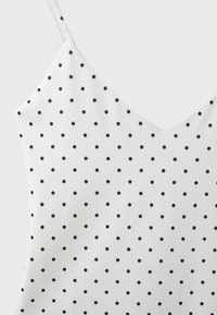 Haut blanc sans manches avec fines bretelles et petits pois noirs uniformément espacés, sur fond clair uni.