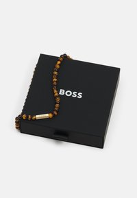 Perlearmbånd lavet af brune og gule tigerefuglestene, med et sølv rektangulært mærke med teksten "BOSS," præsenteret på en sort kasse.