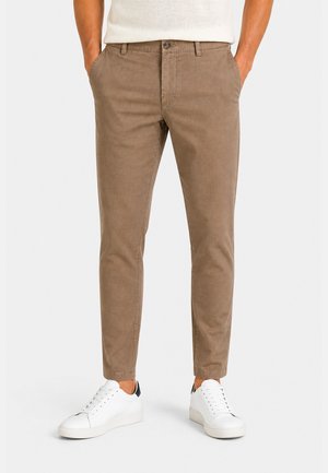 Mann trägt braune Slim-Fit-Hose und weiße Sneaker mit navyblauem Fersen-Detail, Hände in den Taschen, vor weißem Hintergrund.