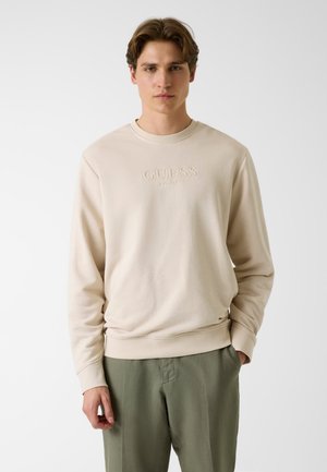 Młody mężczyzna w beżowej bluzy z okrągłym dekoltem z wypukłym logo "GUESS" oraz oliwkowozielonych spodniach, stoi na tle jednolitego białego tła.