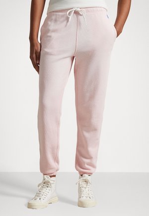 Persoon draagt lichtroze sweatpants met trekkoord en witte hoge sneakers, staand met handen in de zakken tegen een effen achtergrond.