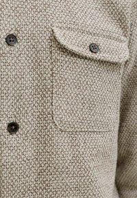 Tissu beige texturé avec un motif tissé, présentant une poche à rabat boutonnée et des boutons brun foncé à quatre trous sur un vêtement.