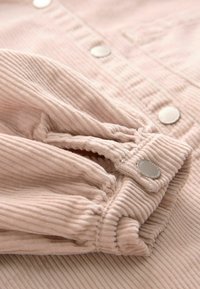 Tejido de sarga de color rosa claro con textura estriada, que presenta mangas fruncidas y botones a presión metálicos. El detalle en primer plano muestra la costura.