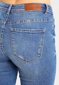 Sininen farkkuhaalari, takatasku ja ruskea nahkakappale vyötäröllä. Kevyesti kuluneet pinnat ja näkyvät ompelusaumat.