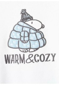 Vyšívaná plyšová tkanina zobrazuje kreslené zviera v modrom prikrývke s bielymi hviezdami, ktoré má na sebe sivú pletenú čiapku. Text znie "TEPLÉ A PRIEMNÉ."