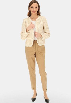 Kobieta ubrana w kremową kurtkę o fakturze, biały top, beżowe spodnie z paskiem ściągającym oraz czarne spiczaste baleriny, stojąca na jednolitym tle.