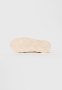 Semelle d'une chaussure beige avec un motif de semelle ondulée, présentée sur un fond blanc uni.