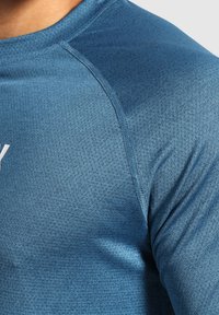Camiseta deportiva de manga corta en tejido azul texturizado. Cuenta con un cuello redondo, detalles de costura en contraste y un logo blanco en el frente.