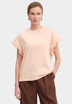Mujer que lleva una camiseta de manga corta rosa claro con mangas de encaje con volantes y pantalones marrones de cintura alta, de pie con una mano en el bolsillo.