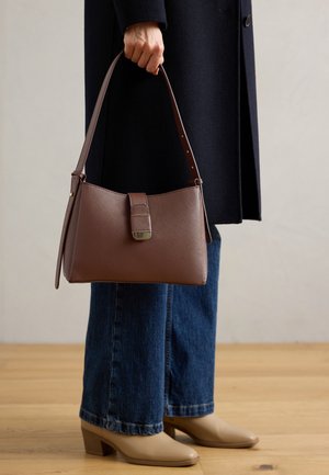 Une main tenant un sac à main en cuir marron avec boucle, portant un manteau sombre, un jean bleu et des chaussures à talons beige sur un sol en bois.