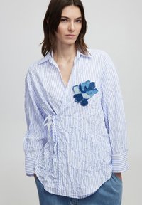 Blusa bianca e azzurro chiaro a righe con un'applicazione floreale in denim sul petto, caratterizzata da un dettaglio legato in vita e una texture stropicciata.