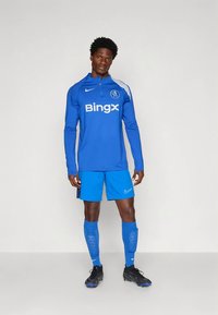 Blauw sportief shirt met lange mouwen en een ritskraag, wit logo, bijpassende blauwe korte broek en zwarte voetbalschoenen. Accent: blauwe sokken en scheenbeschermers.