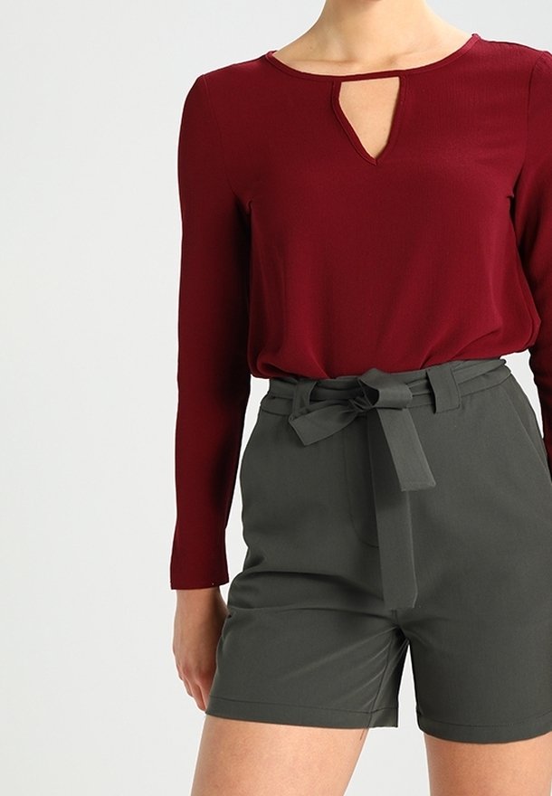 Femme portant un haut bordeaux à manches longues avec un col clé et un short taille haute vert foncé noué avec une ceinture en tissu.