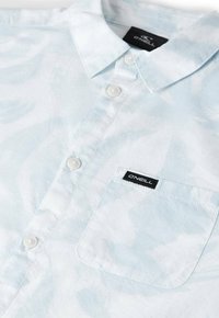 Camisa de color azul claro con botones, hecha de tela con un patrón sutil. Cuenta con cuello, bolsillo delantero y una etiqueta de marca negra.