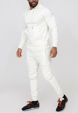 Homme debout portant un sweat à capuche tout blanc et un pantalon cargo jogger avec des baskets noires et orange sur un fond gris uni.